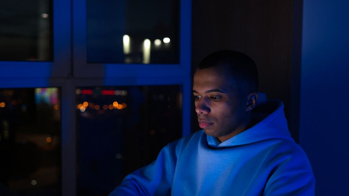 A person works on a laptop at night, lit by the screen, in front of a dark city window.
